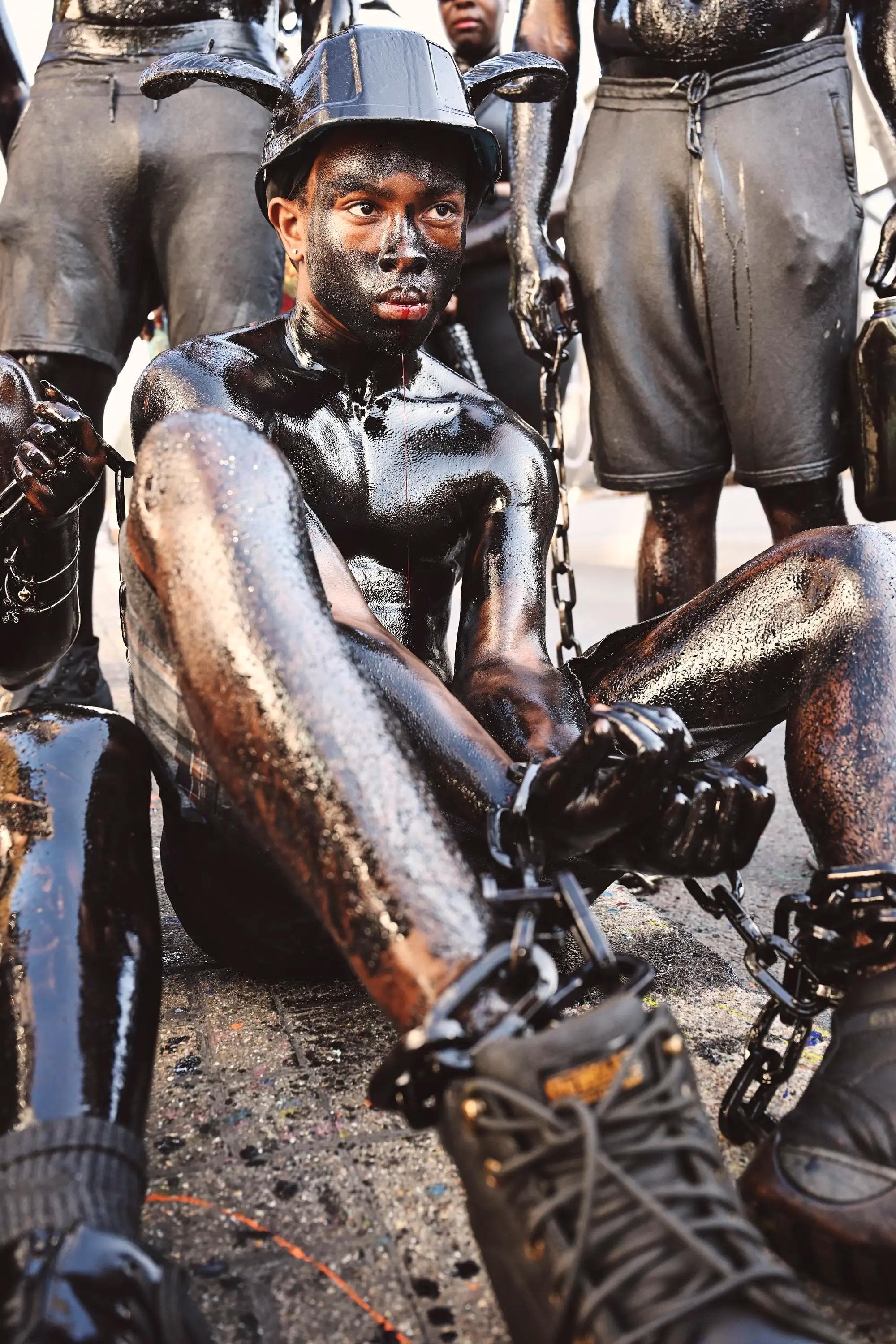 young Jab Jab performer sitting on pavement in black body paint and chains during carnival in London