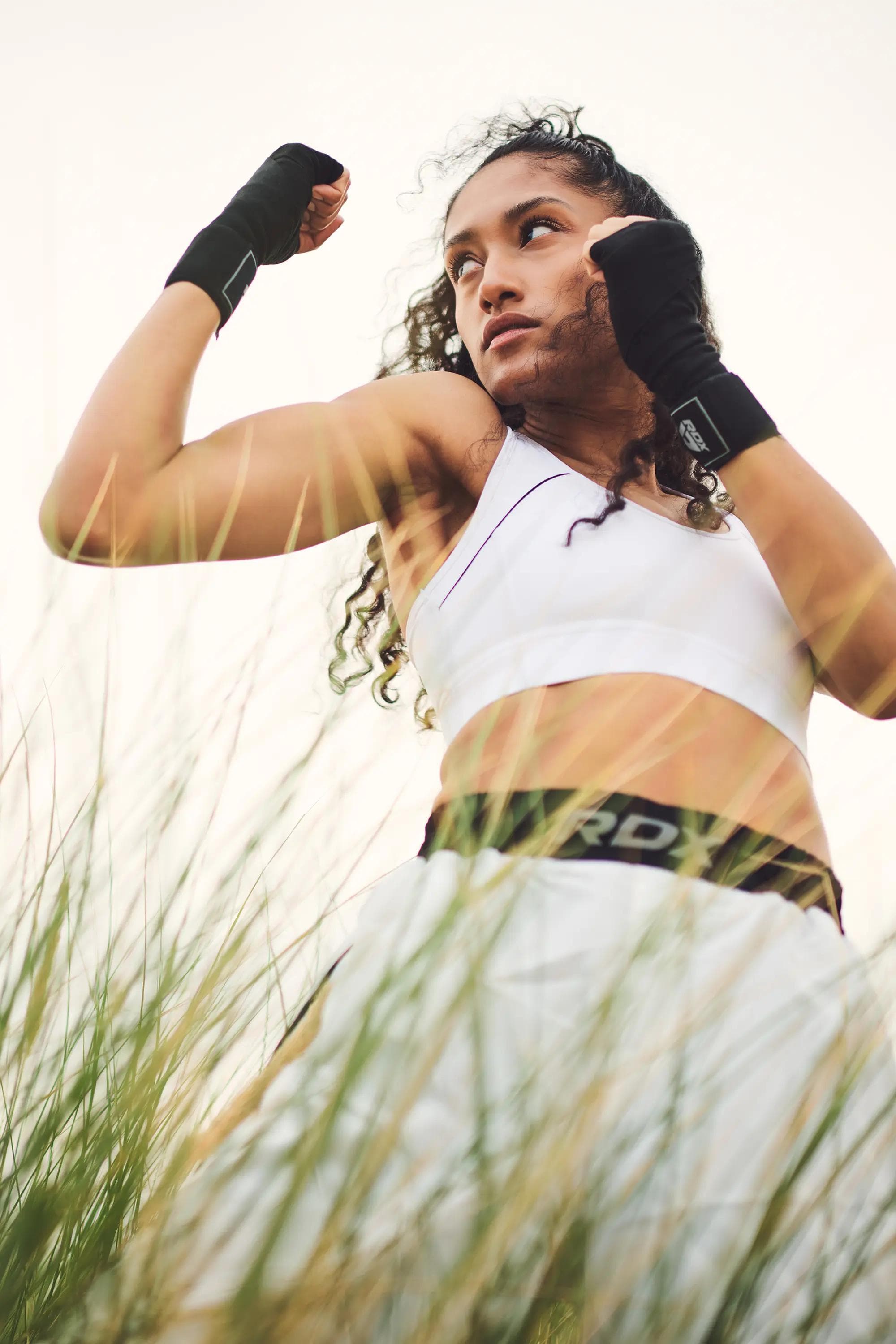 low angle portrait of female combat athlete in RDX training apparel with boxing hand wraps raised in guard position among beach grass