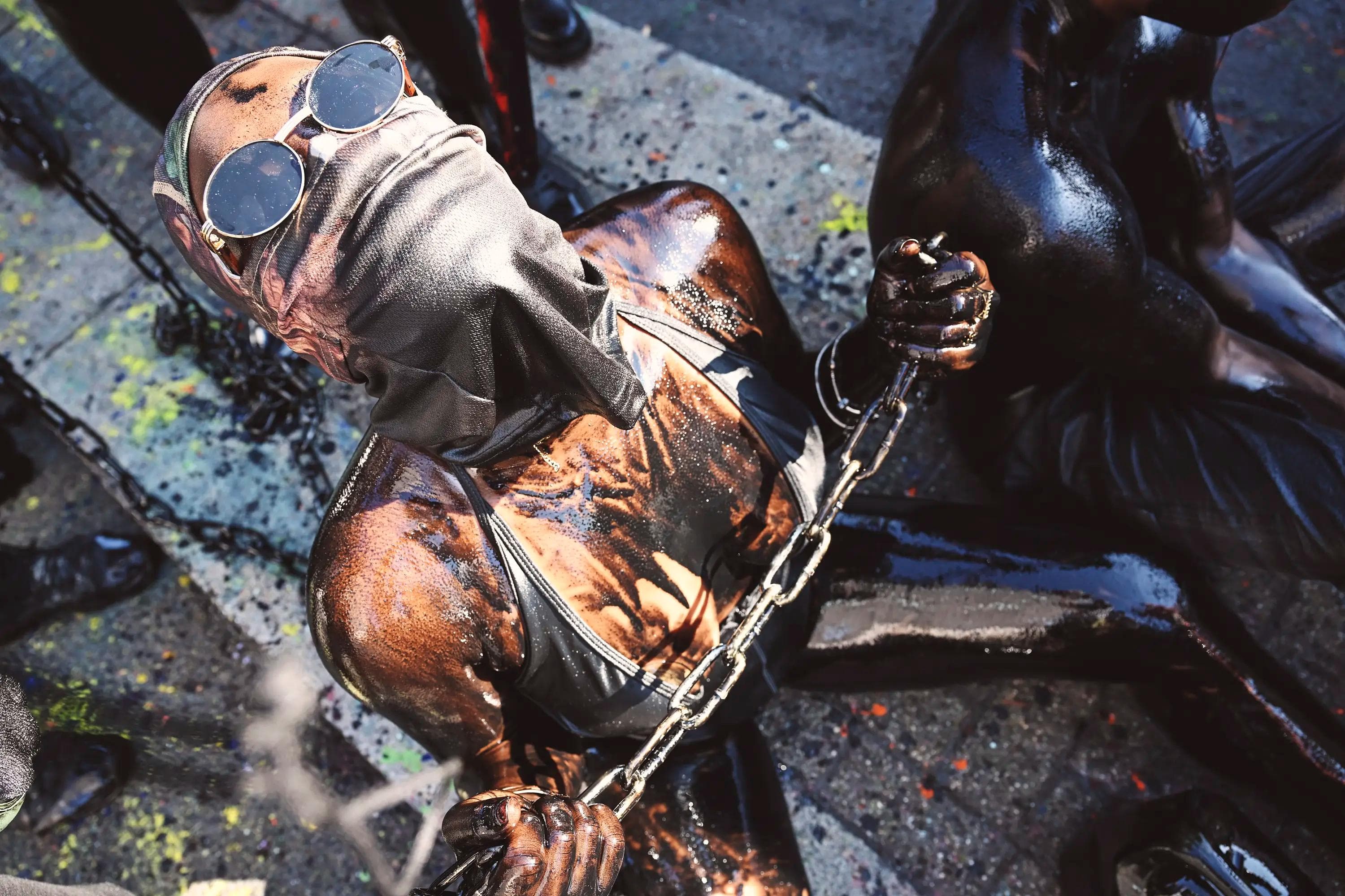 masked Jab Jab carnival participant covered in oil paint and holding chains during street performance