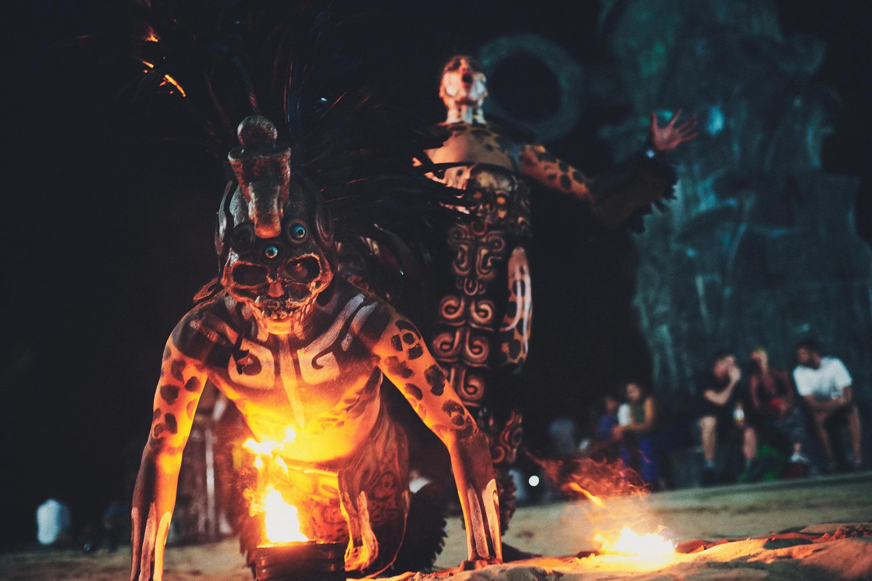 fire performer crawling beside flames while another painted performer stands in the background during a ritual reenactment