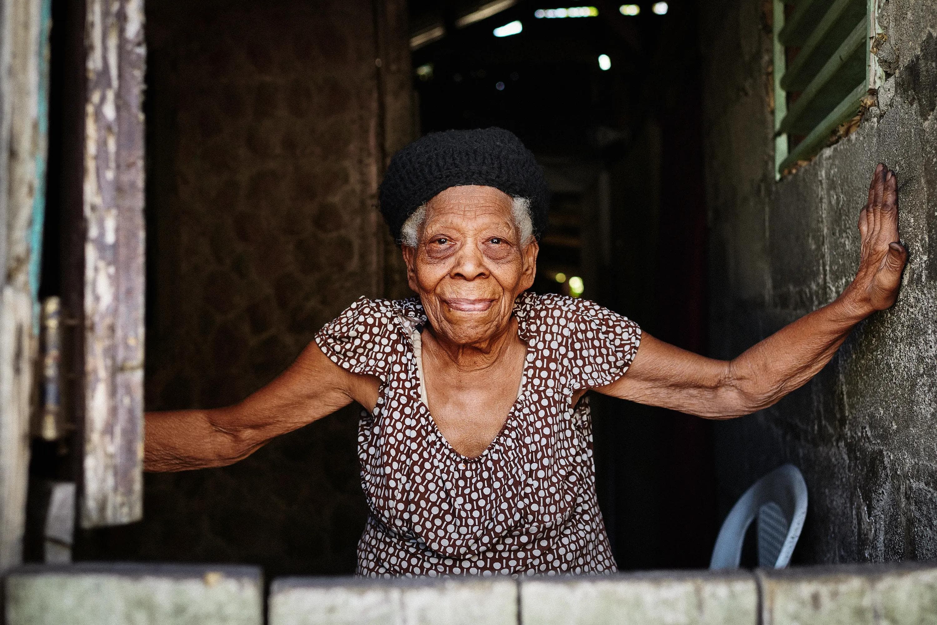 elderly woman standing in dark doorway with arms extended, intimate environmental portrait Dominican Republic
