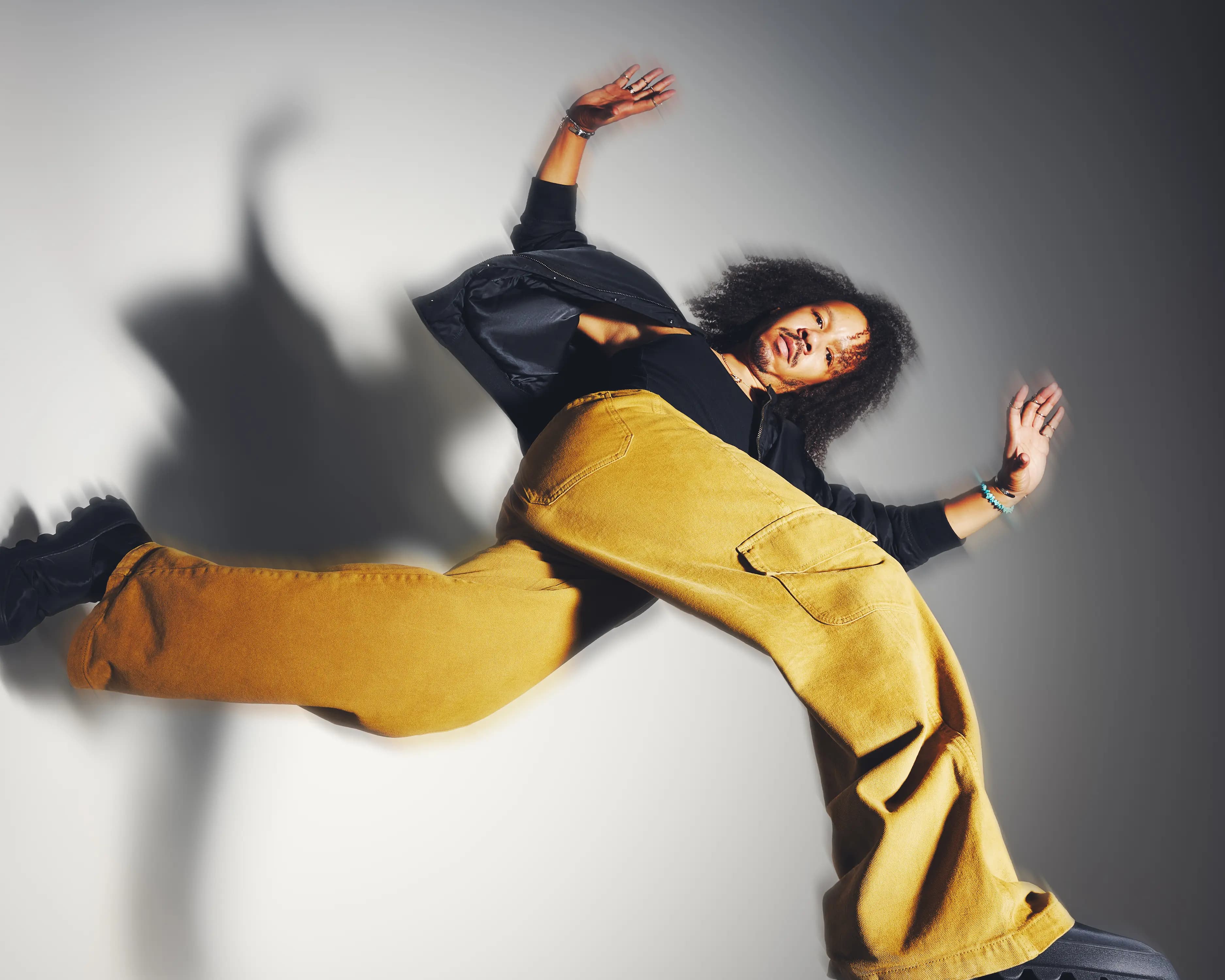 mannequin homme en veste en satin rose et pantalon large jaune assis sur un tabouret, effet de traînée lumineuse en mouvement, photographie de mode éditoriale à Londres
