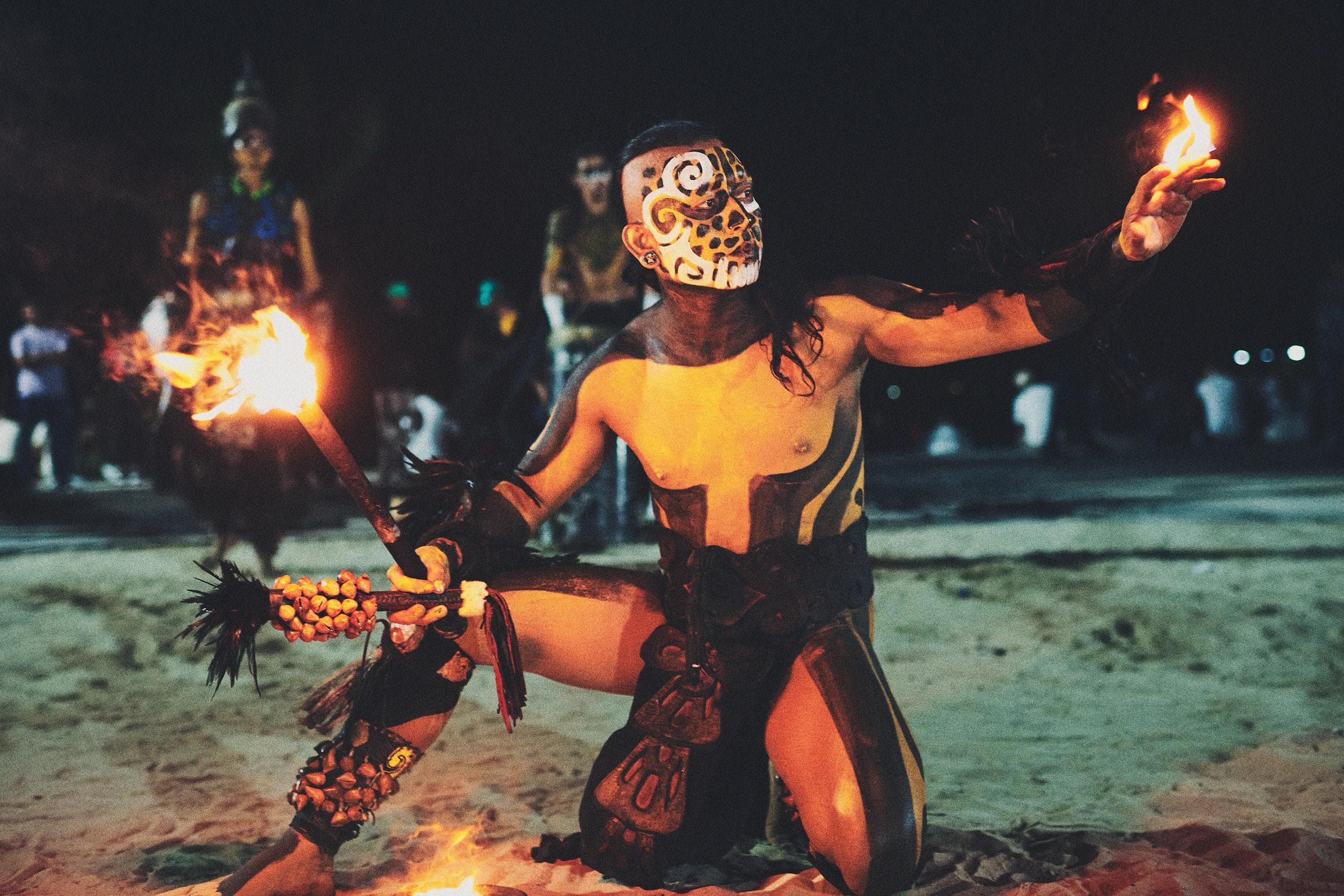 performer with ceremonial body paint and torch extending a hand toward fire during a nighttime beach performance