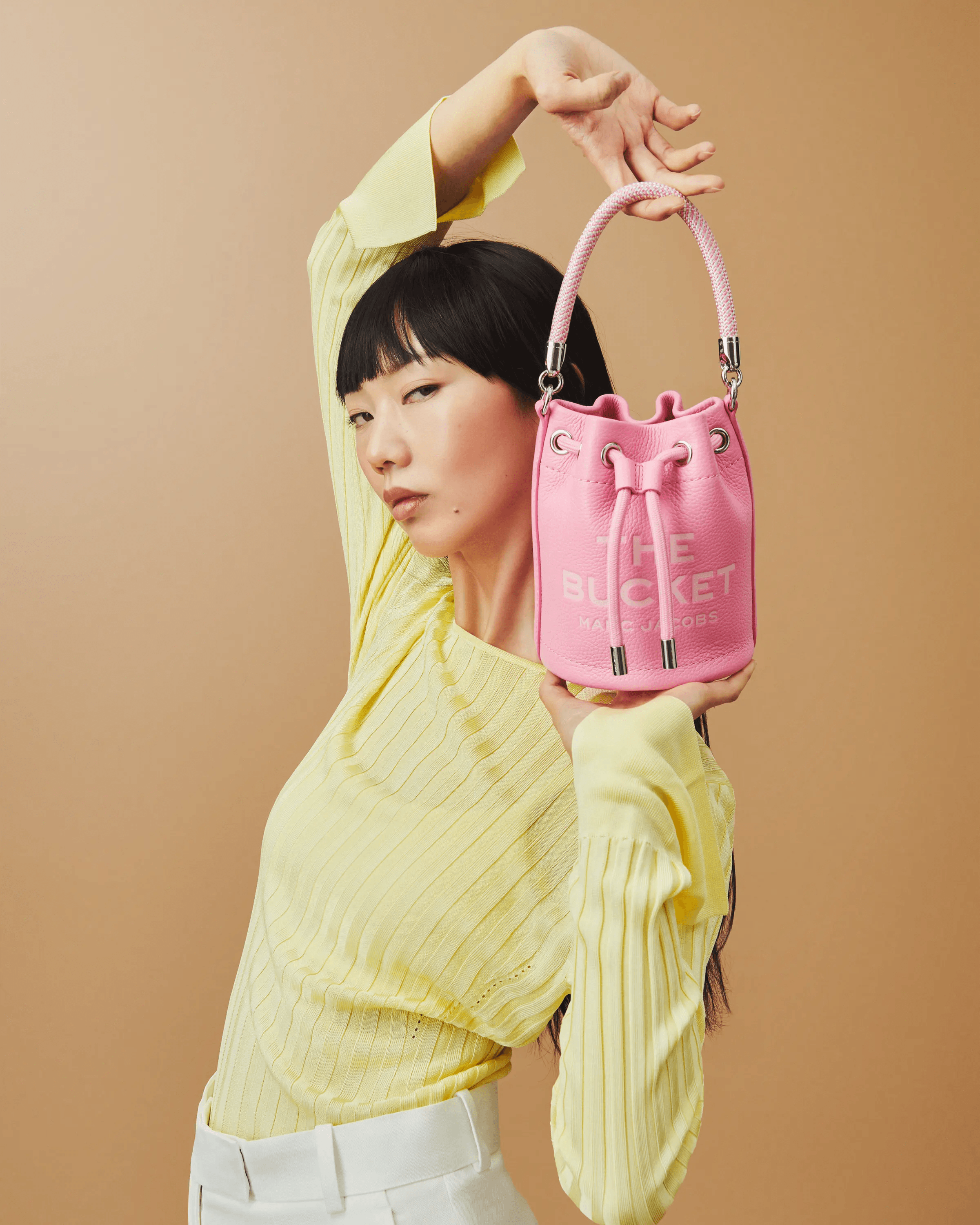 female model in yellow knit raising arm with pink Marc Jacobs The Bucket bag, dynamic studio composition, premium fashion campaign London