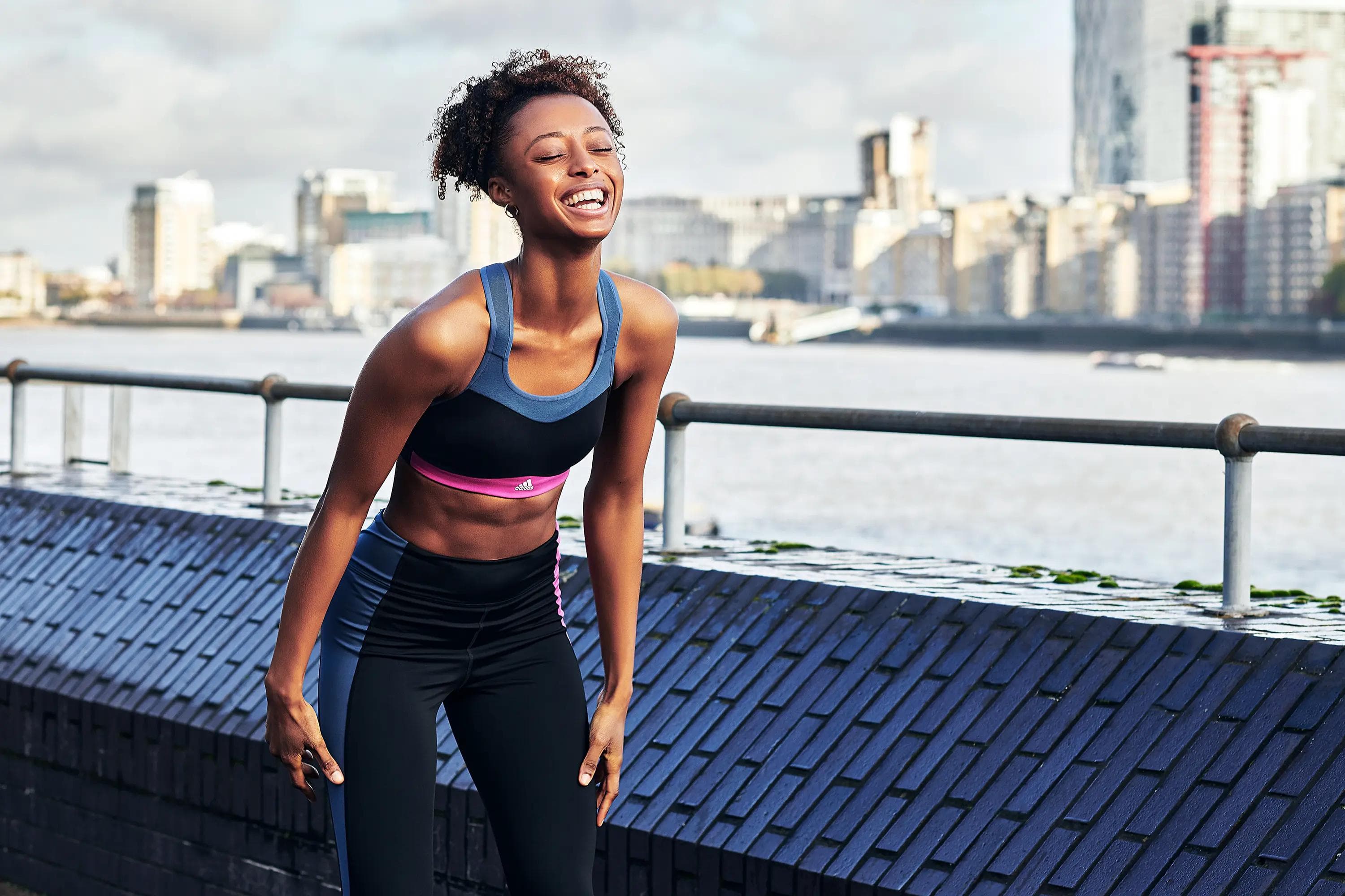female athlete smiling beside riverside railing with city skyline in background, lifestyle fitness photography London